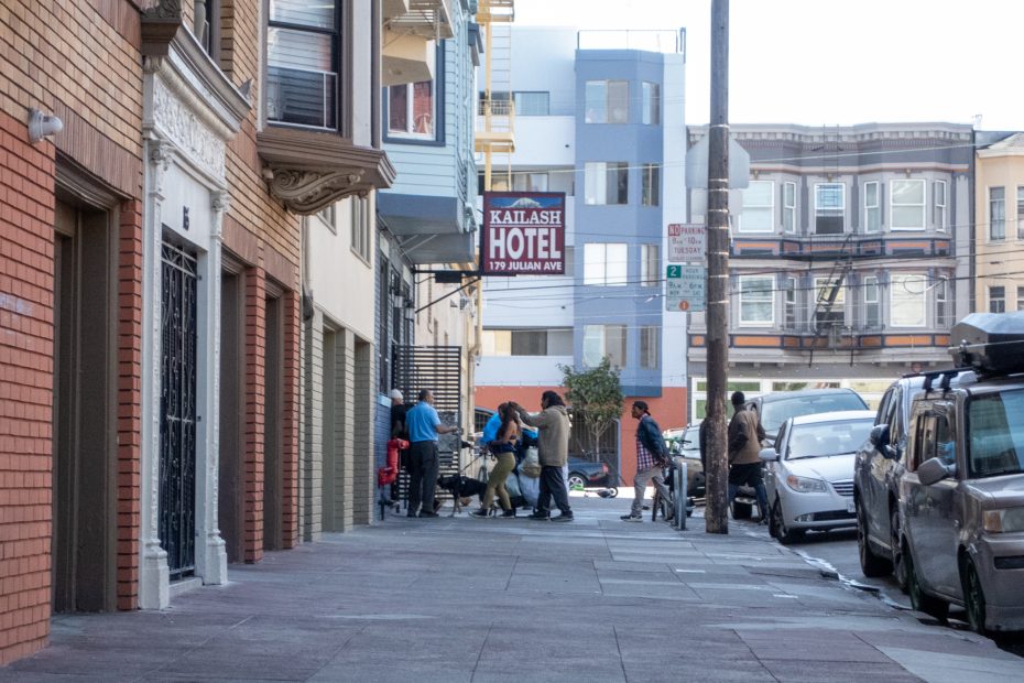 People interacting and standing on an urban sidewalk near a hotel sign and parked cars. Buildings and utility poles line the street.