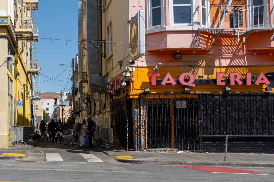 Narrow urban alley with people and dogs. Tall buildings on both sides. Pink building with "TAQUERIA" sign on the right. Sunny day with casts of shadows on the street.