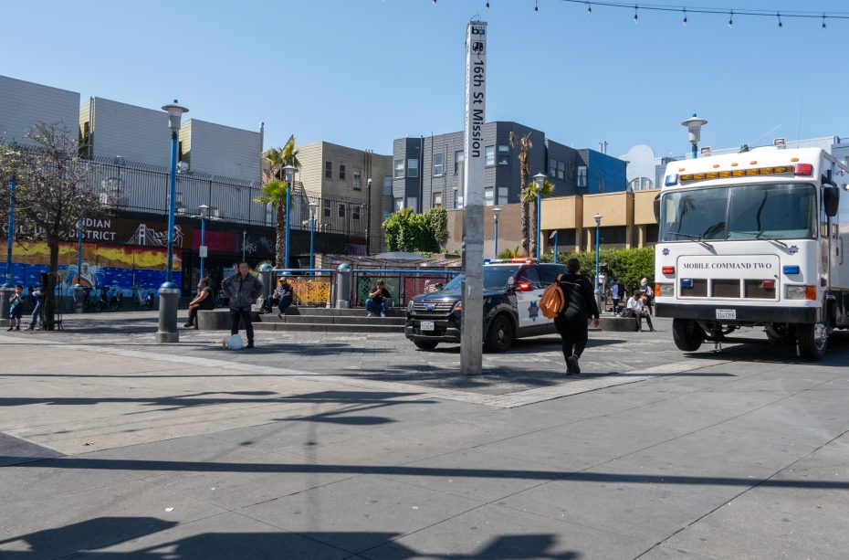 City street scene with an emergency vehicle and police car parked. People walk nearby; buildings and a colorful mural in the background. Sunny day.