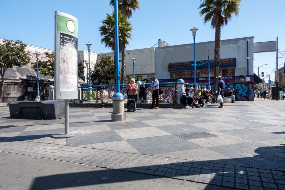 Urban scene with people sitting and standing in a plaza. Palm trees line the area, and a subway entrance is in the background. A colorful mural is painted on a building nearby.