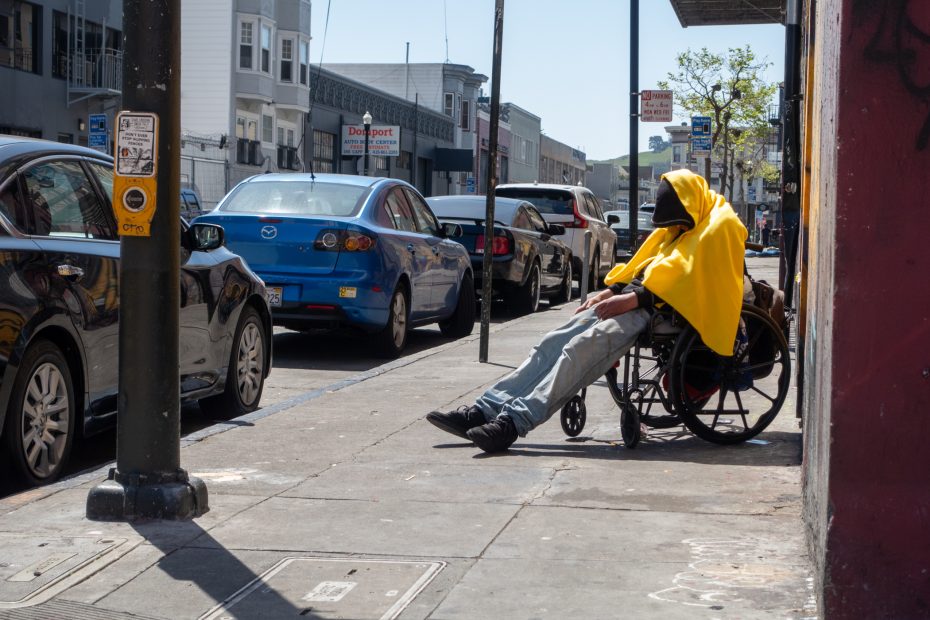 Person in a yellow hoodie sits in a wheelchair on a city sidewalk, facing the street. Cars are parked along the road in an urban setting.