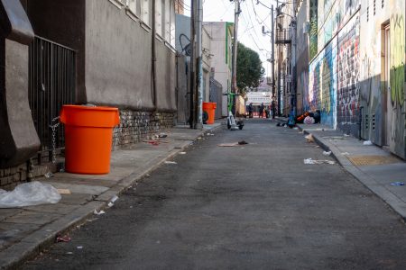 A narrow, graffiti-filled alley with scattered trash and orange bins lining the walls. People are visible in the distance, and utility lines crisscross above.