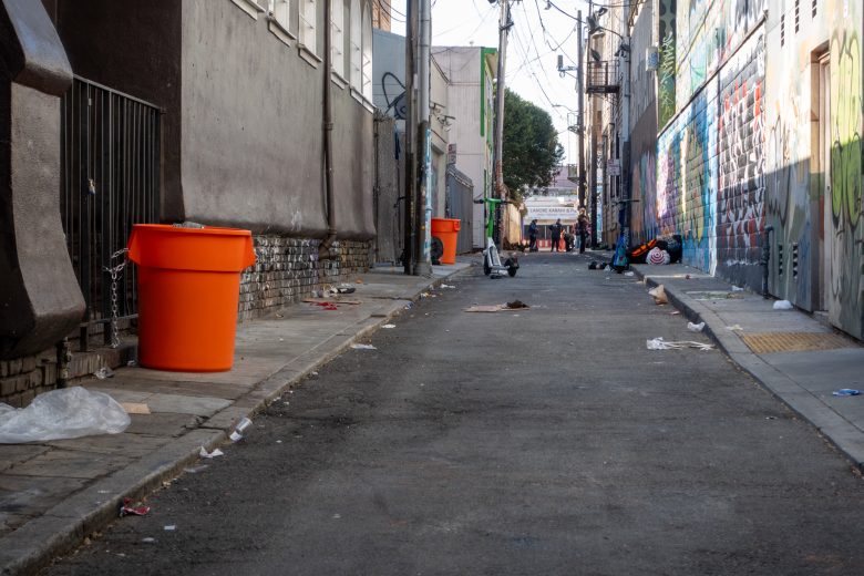 A narrow, graffiti-filled alley with scattered trash and orange bins lining the walls. People are visible in the distance, and utility lines crisscross above.