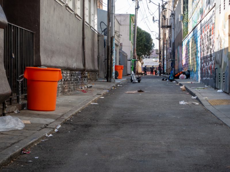A narrow, graffiti-filled alley with scattered trash and orange bins lining the walls. People are visible in the distance, and utility lines crisscross above.