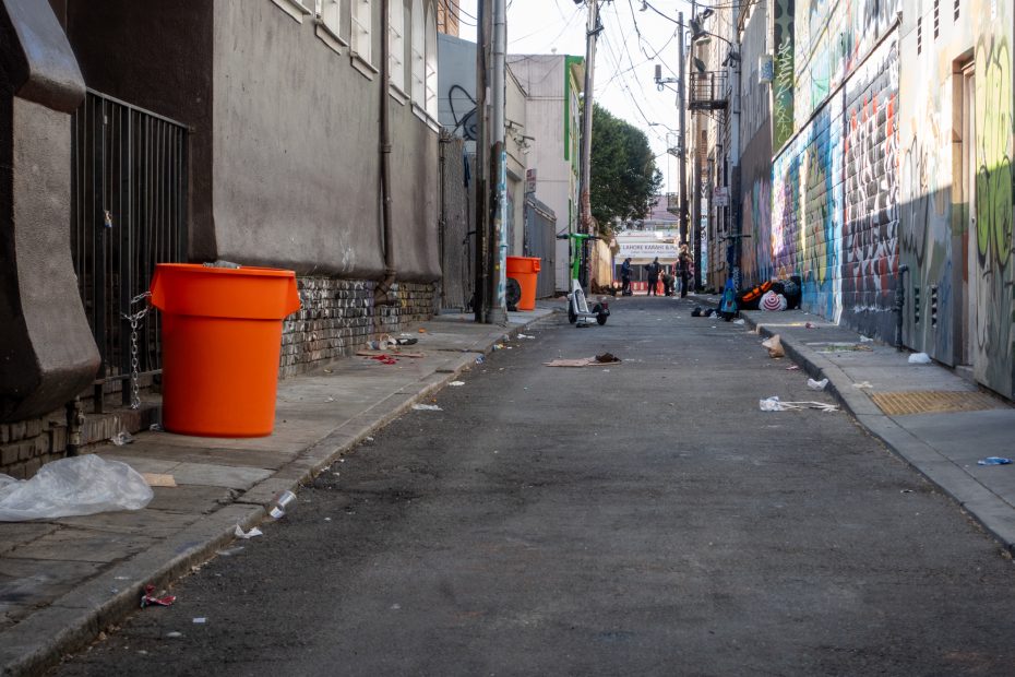 A narrow, graffiti-filled alley with scattered trash and orange bins lining the walls. People are visible in the distance, and utility lines crisscross above.