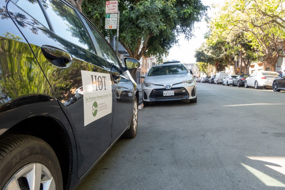 Two parked cars on a sunny street, one with a San Francisco Homeless Outreach vehicle sign. Trees and additional parked cars line the street in the background.
