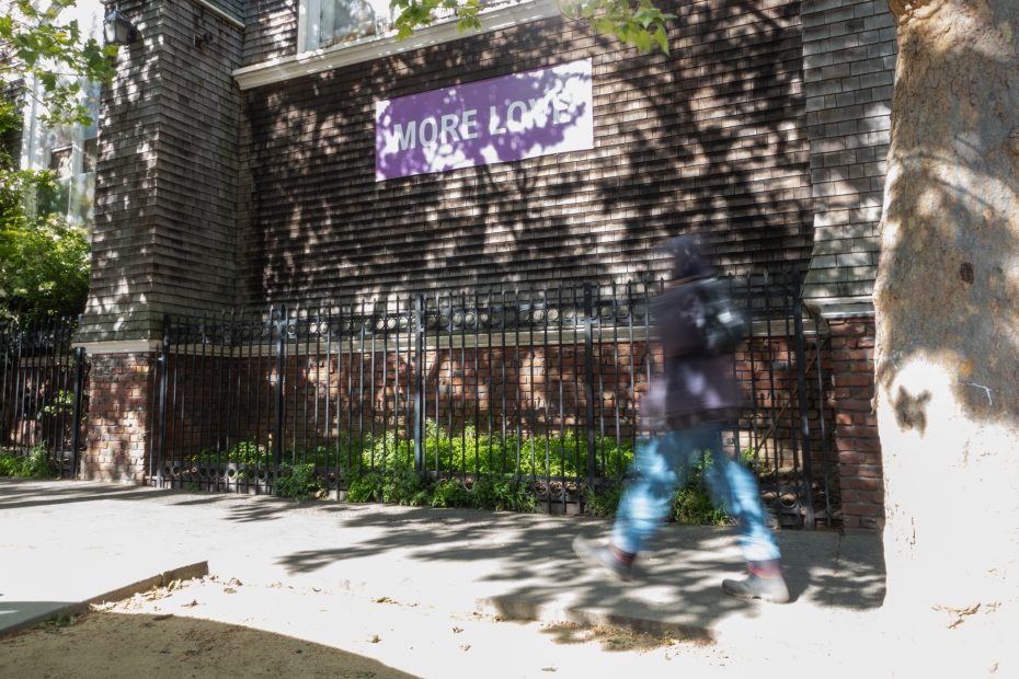 A person walks past a brick building with a purple sign that reads "MORE LOVE." Shadows from trees cover the sidewalk.
