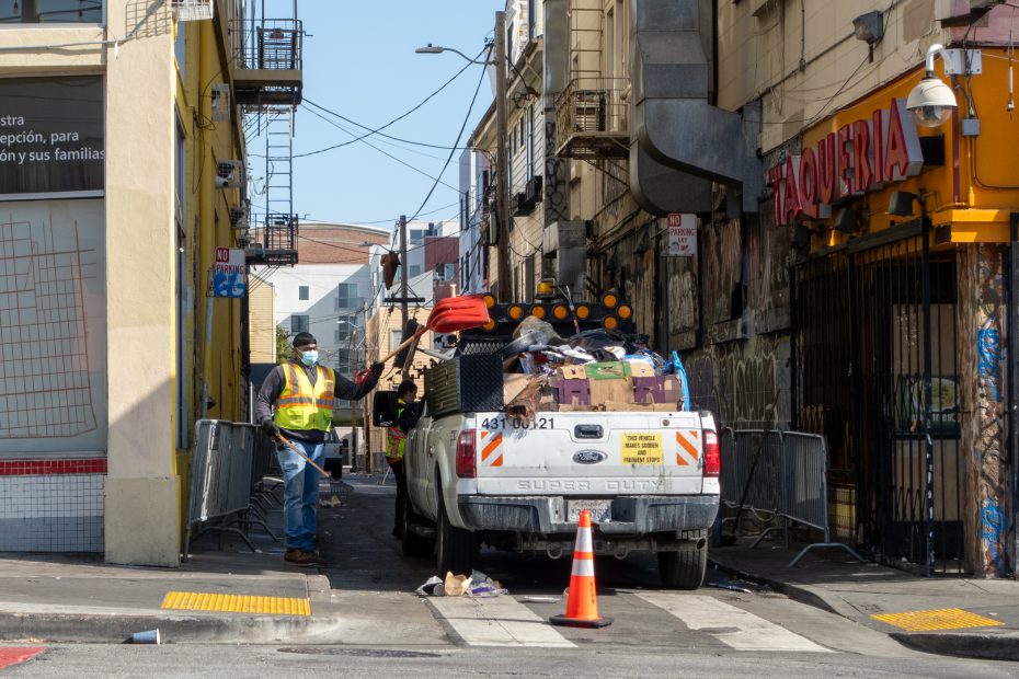 A worker wearing a safety vest and mask stands beside a white utility truck loaded with items in a narrow urban alley. Orange safety cones are placed near the truck's rear.