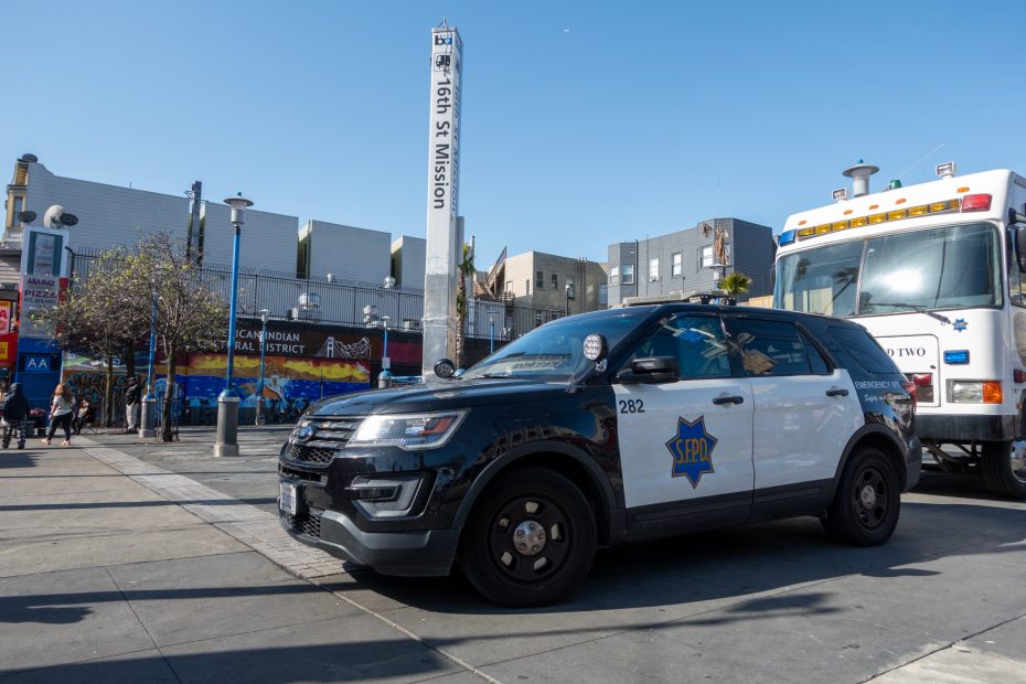 Police SUV parked in an urban area next to a building with a sign reading "Cambodian Cultural District" and a street post indicating the intersection of 16th St and Mission.