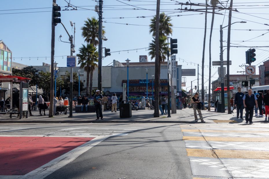 A busy urban intersection with people crossing the street, surrounded by palm trees and buildings. Traffic lights and overhead wires are visible under a clear blue sky.