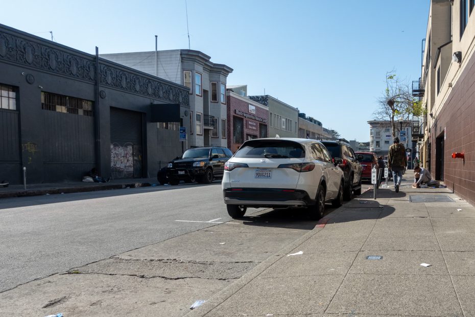 Street view with parked cars, a police car, and people walking and sitting near a building. A few trees and scattered litter are visible on the sidewalk under a clear sky.