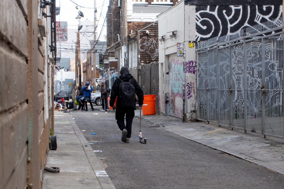 A person with a backpack walks a scooter down a graffiti-covered alley, while several people gather near trash and bags in the distance.