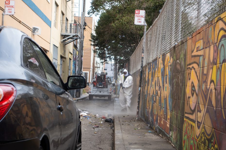 A person in protective gear power washes a littered alleyway next to a graffiti-covered wall, while a black car is parked nearby.