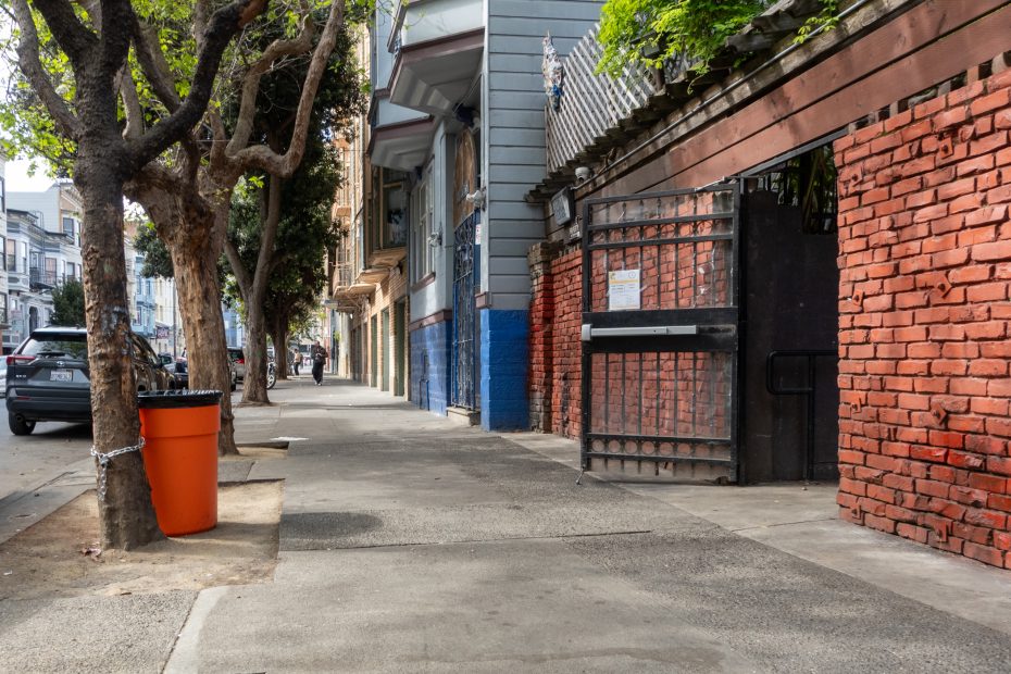A city sidewalk with trees, a red brick wall, an open black metal gate, and an orange trash bin chained to a tree. Cars and buildings line the street in the background.