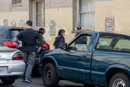 Several people stand on a street near parked cars, including a man wearing a vest and another person in a police hat, in front of a brick building with decorative tiles on the wall.