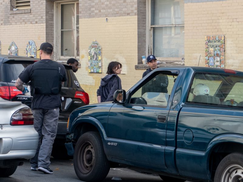 Several people stand on a street near parked cars, including a man wearing a vest and another person in a police hat, in front of a brick building with decorative tiles on the wall.