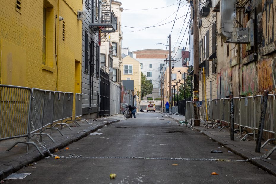 A narrow urban alley lined with metal barricades, scattered trash, and graffiti on walls. A person with a bicycle is visible in the distance.