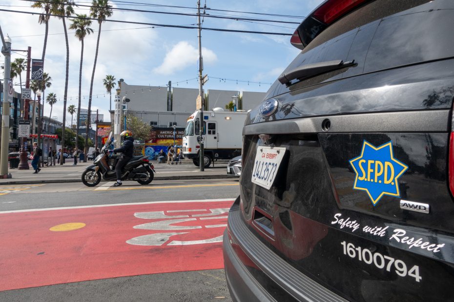 A San Francisco police vehicle is parked on a city street near a bus lane, with a police motorcycle and van visible in the background.