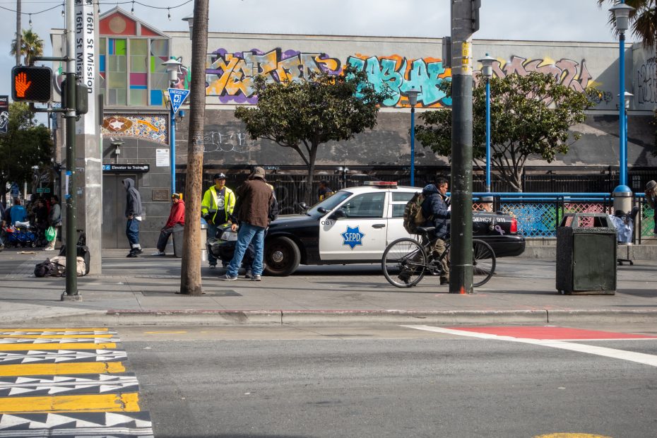 A police car is parked on a city street as officers and pedestrians stand nearby; graffiti covers the building in the background.