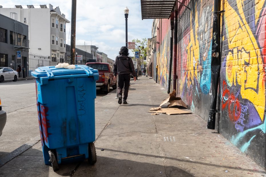 A person walks down a city sidewalk lined with colorful graffiti and a blue trash bin in the foreground. Cardboard and parked cars are visible along the street.