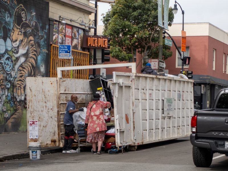 Two people search through a large white dumpster on a city street next to a mural and a parked truck, with buildings and signs visible in the background.