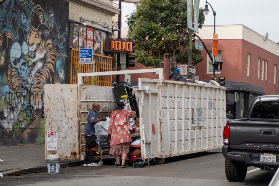 Two people search through a large white dumpster on a city street next to a mural and a parked truck, with buildings and signs visible in the background.