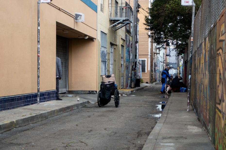 A narrow urban alleyway with a cart in the center, two people near bags on the right, and another person partly visible on the left by a building.