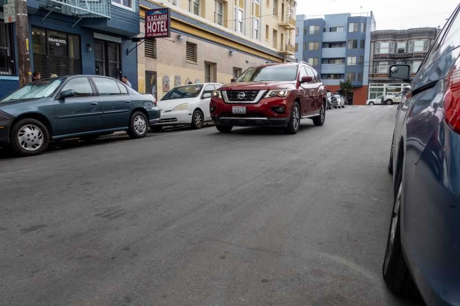 A red SUV drives up a city street lined with parked cars, buildings, and a hotel sign on the left side.