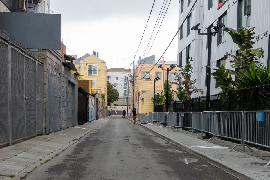 A narrow urban alley lined with fences and modern buildings, two people walking in the distance, and metal barricades on the right side.