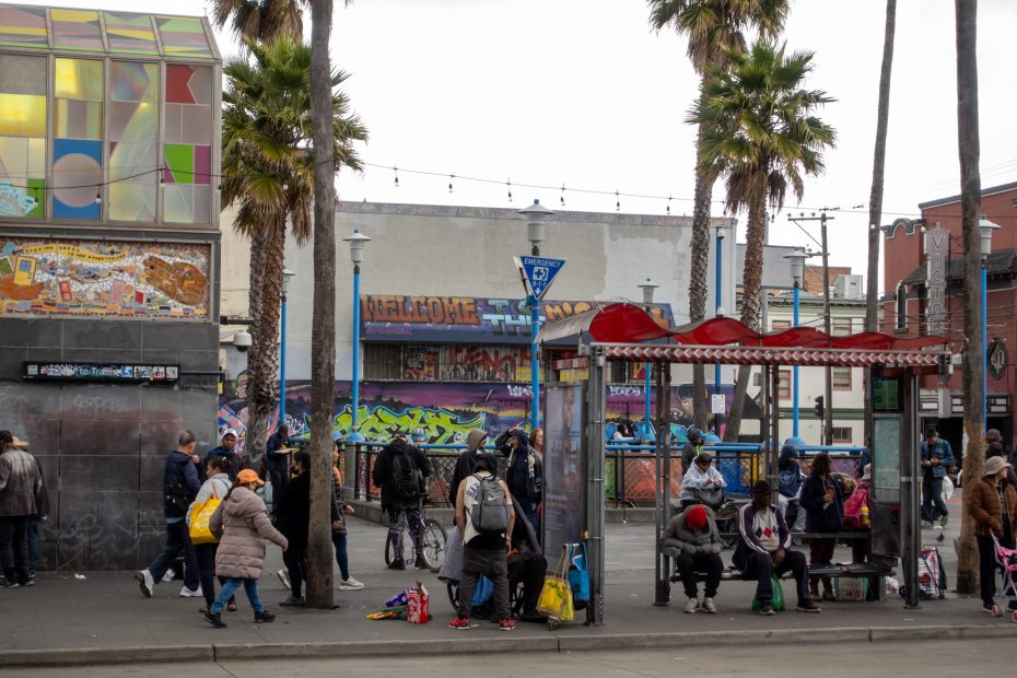 People gather and sit near a bus stop in an urban area with palm trees, colorful murals, and a "Welcome" sign in the background.