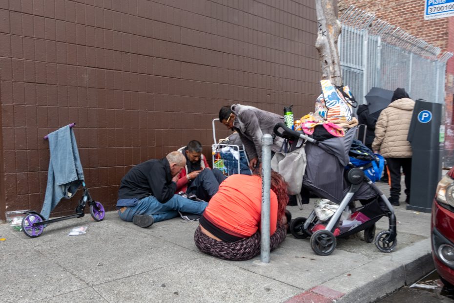 Several people are sitting and crouching on a sidewalk beside a wall, surrounded by personal belongings and a stroller loaded with items.