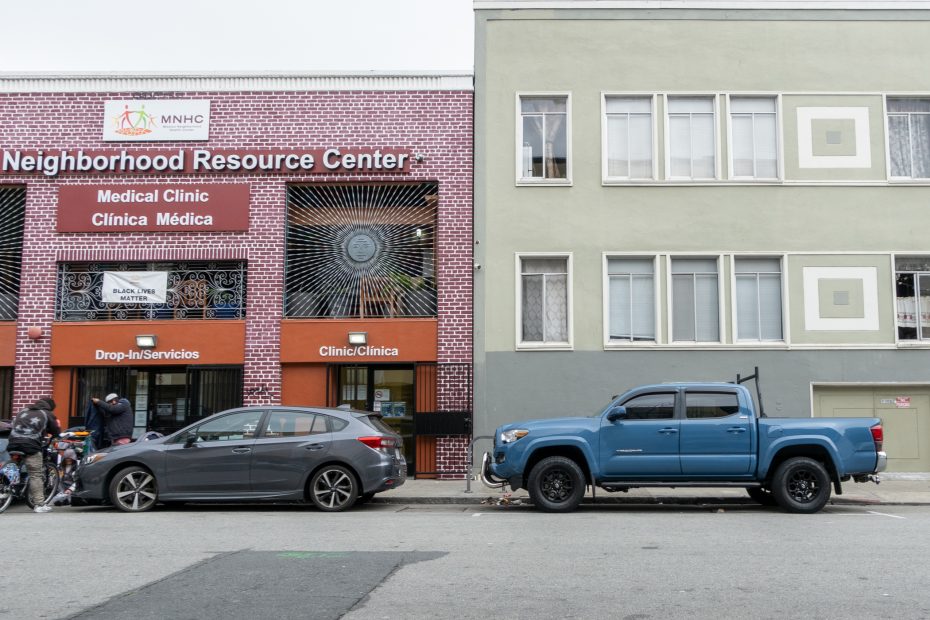 Two cars are parked on the street in front of a Neighborhood Resource Center with a Medical Clinic; three people stand near bicycles on the sidewalk.