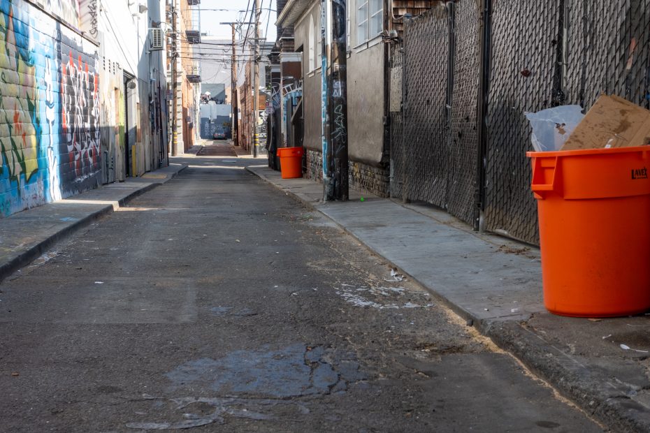 A narrow urban alleyway with graffiti on one wall, chain-link fencing on the other, and two large orange trash bins near the sidewalk.