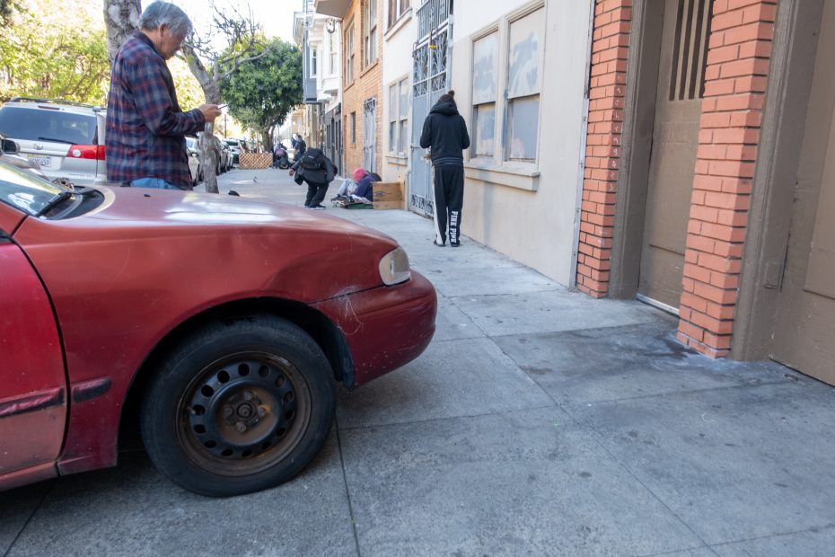 A man stands next to a red car with a dented hood, while another person walks past boarded windows on a city sidewalk.