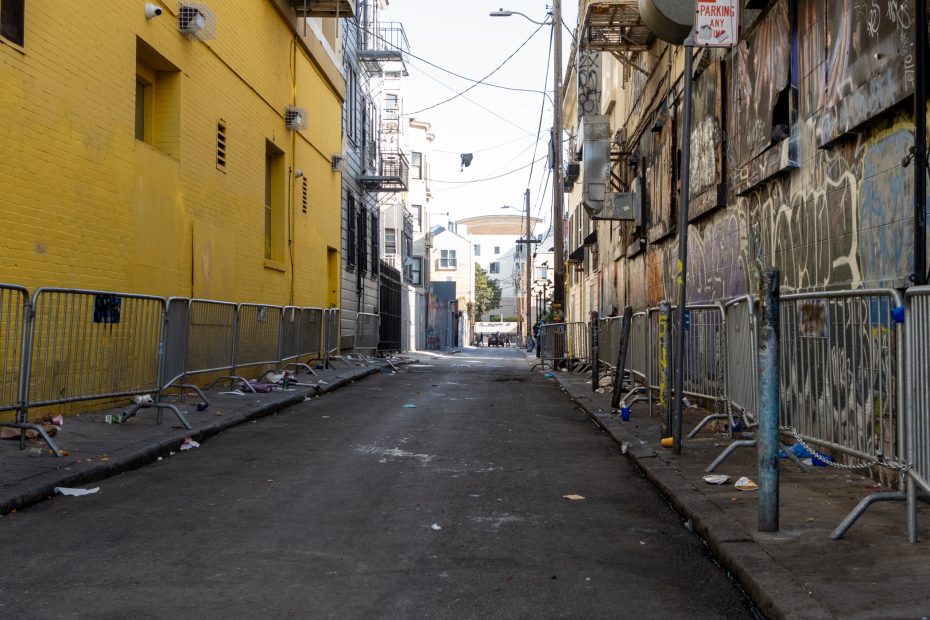 A narrow urban alley lined with metal barricades, graffiti-covered walls, scattered trash, and a yellow building on the left side.
