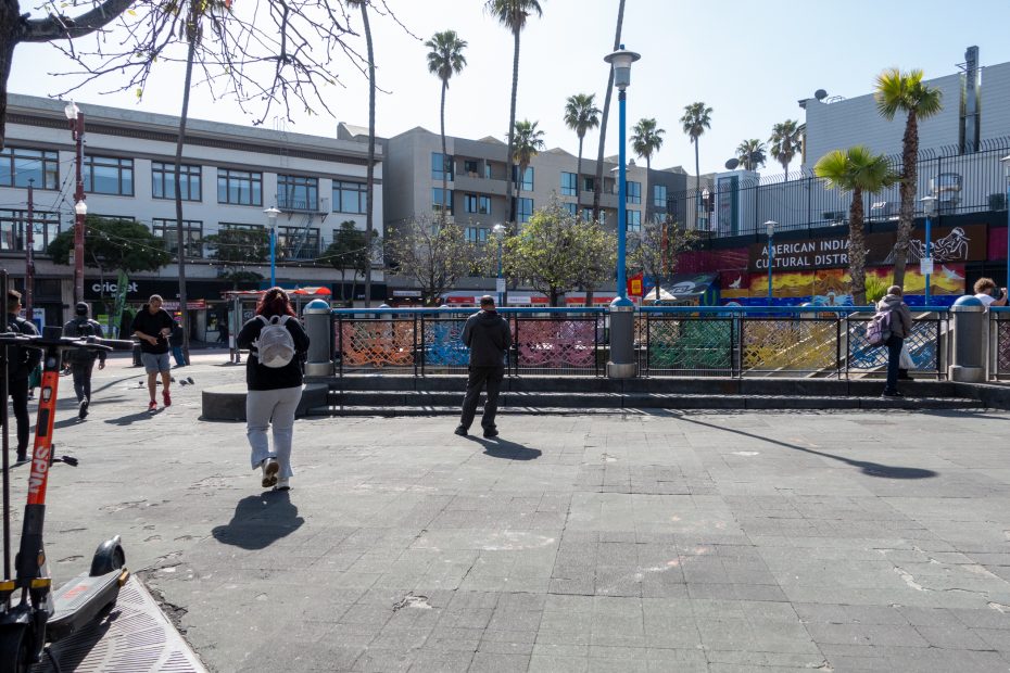 People walk near a fenced area in an urban plaza with palm trees, scooters, and a sign that reads "American Indian Cultural District" in the background.