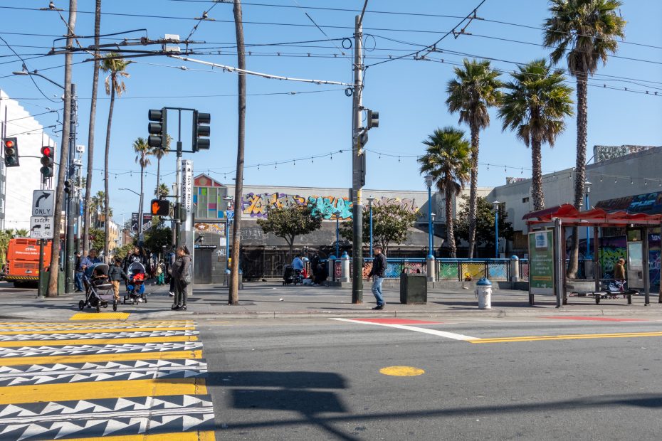 Urban street intersection with palm trees, utility poles, crosswalk, people waiting, and buildings featuring colorful murals under a clear blue sky.