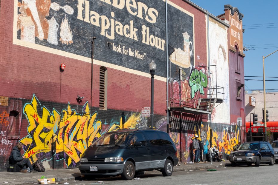 A city street scene with a van and car parked by a building featuring large murals, graffiti, and a faded "flapjack flour" advertisement. Several people are visible on the sidewalk.