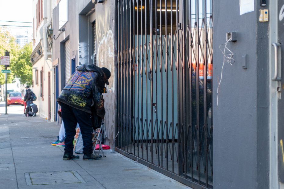 Two people stand on a city sidewalk near a gated storefront, with one person leaning toward the wall and another figure in the background.