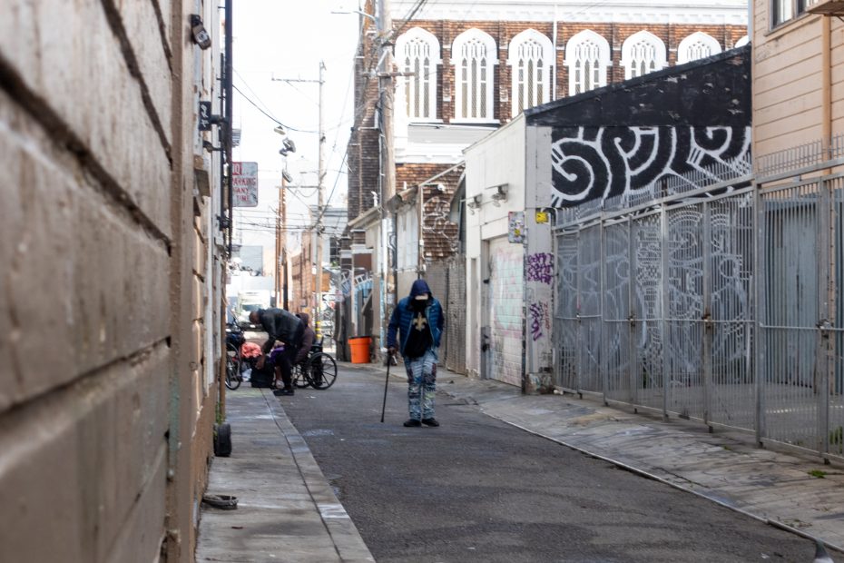 A person in a blue hoodie walks down an urban alleyway while two others attend to a bicycle near a metal fence; buildings line both sides of the alley.