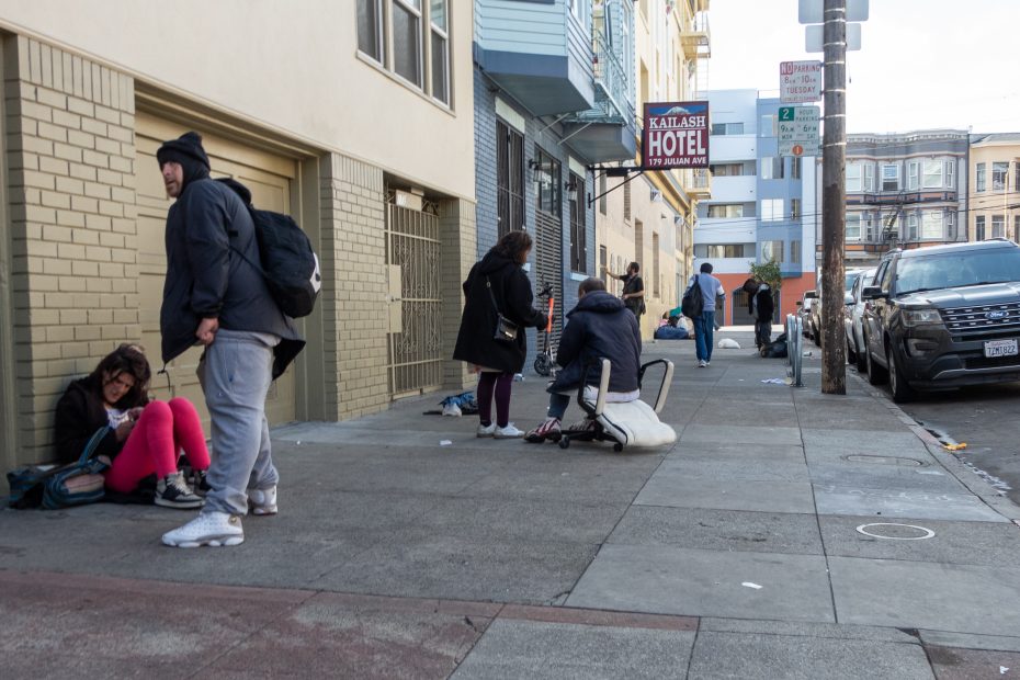 Several people sit and stand on a city sidewalk near buildings and parked cars in front of the Kailash Hotel during daytime.