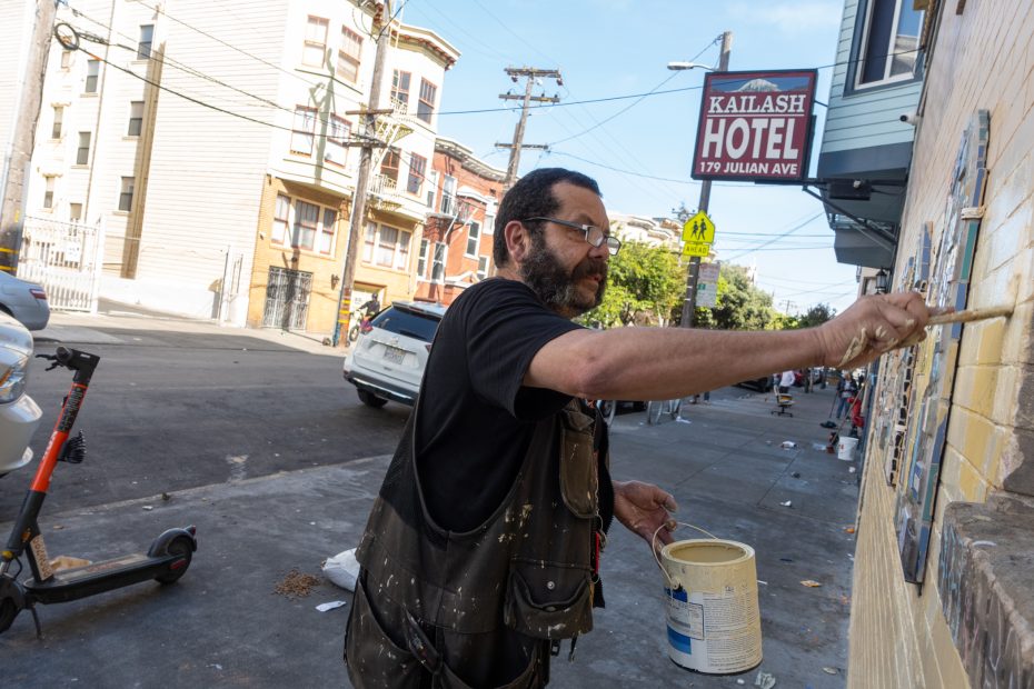 A man paints a mural on an exterior wall along a city street near a hotel sign that reads “Kailash Hotel, 179 Julian Ave.”.