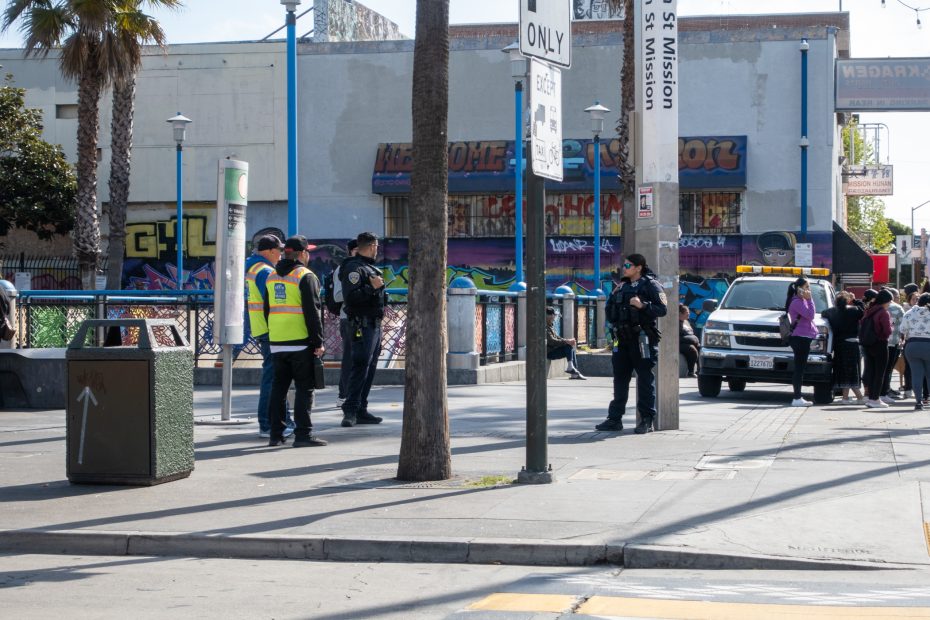 Police officers and a person in a safety vest stand on a city sidewalk near a group of people and a parked police vehicle, with graffiti-covered buildings in the background.