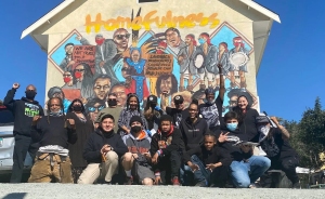 A group of people posing in front of a colorful mural depicting various faces and scenes.