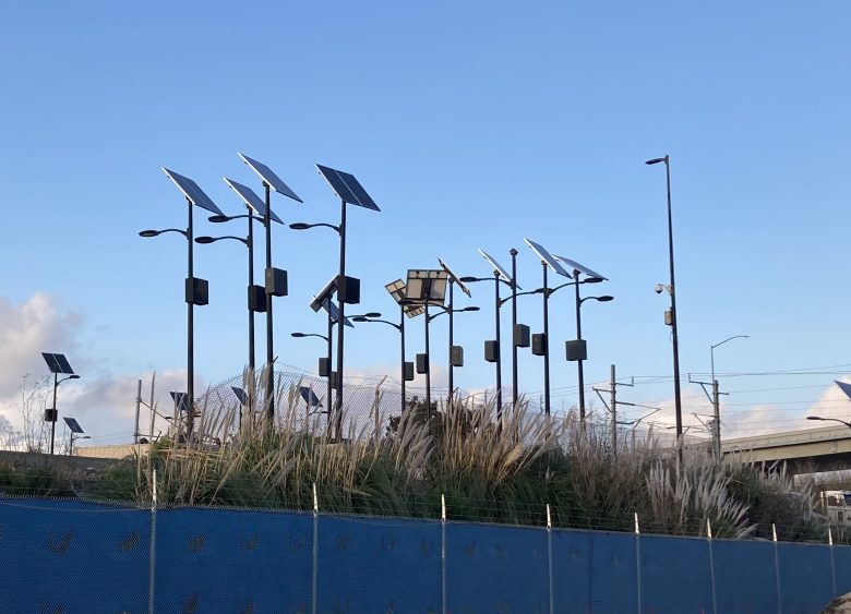 A group of solar-powered streetlights on tall black poles surrounded by vegetation, set against a clear blue sky and highway overpass in the background.
