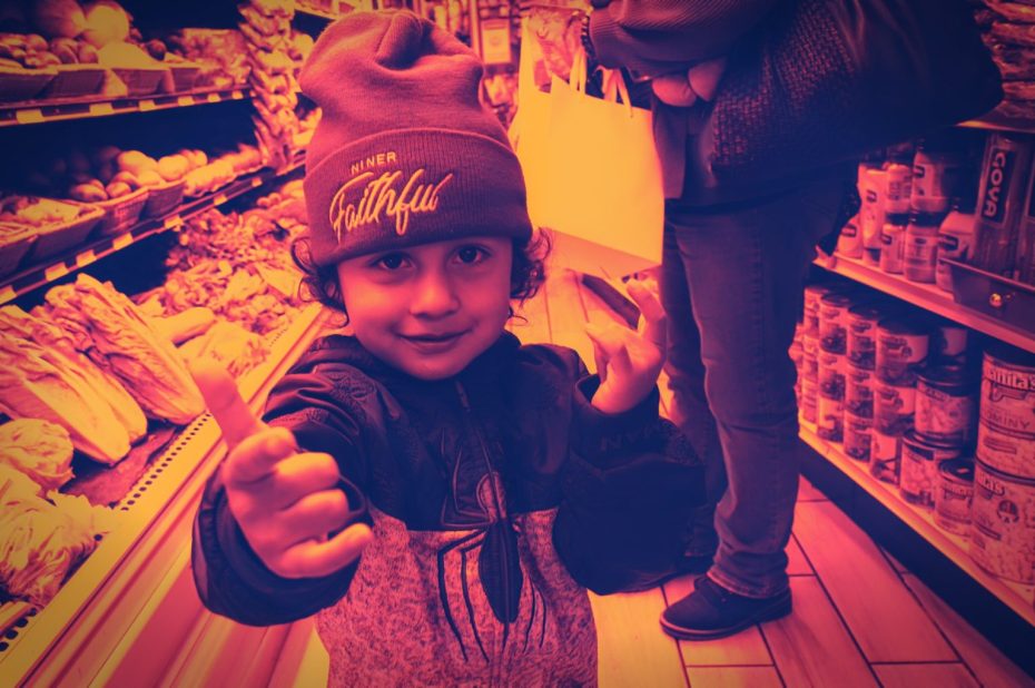 A young child in a beanie gives a thumbs up and points in a grocery store aisle, with shelves of produce and canned goods visible.