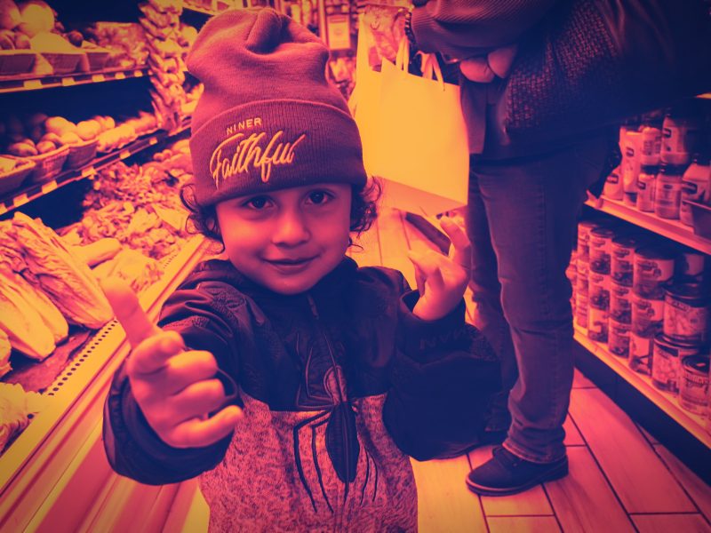 A child wearing a beanie and jacket stands in a grocery store produce aisle, giving a thumbs up while looking at the camera. An adult holding a shopping bag stands nearby.