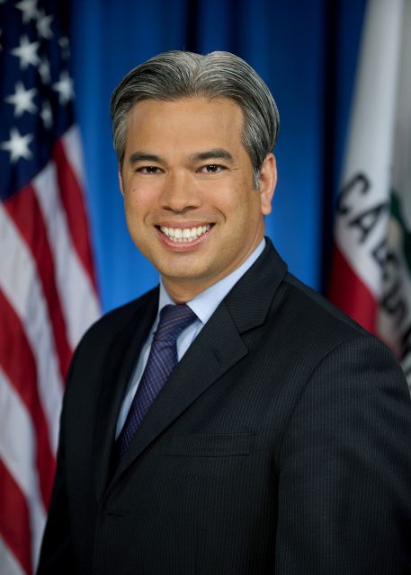 A man in a dark suit and tie smiles in front of American and California state flags against a blue background.