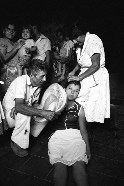 A young woman sits on the floor, supported by a man holding a hat, while a woman touches her head; several people with children stand in the background.