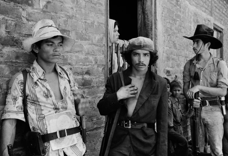 Four men, some armed and in casual clothing, stand near a brick wall in El Salvador as a woman looks out from a doorway and two children stand nearby.